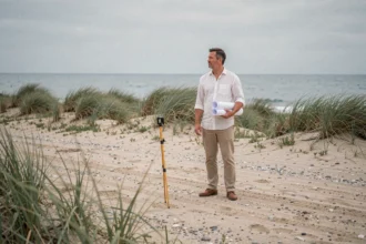 Homme tenant des plans architecturaux sur un terrain en bord de mer à vendre, regardant l'horizon marin